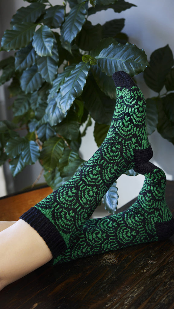 Green and black patterned socks worn by a person sitting on a wooden surface with plants in the background.