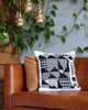 Decorative pillow on a brown couch with greenery and gold pendant lights in the background