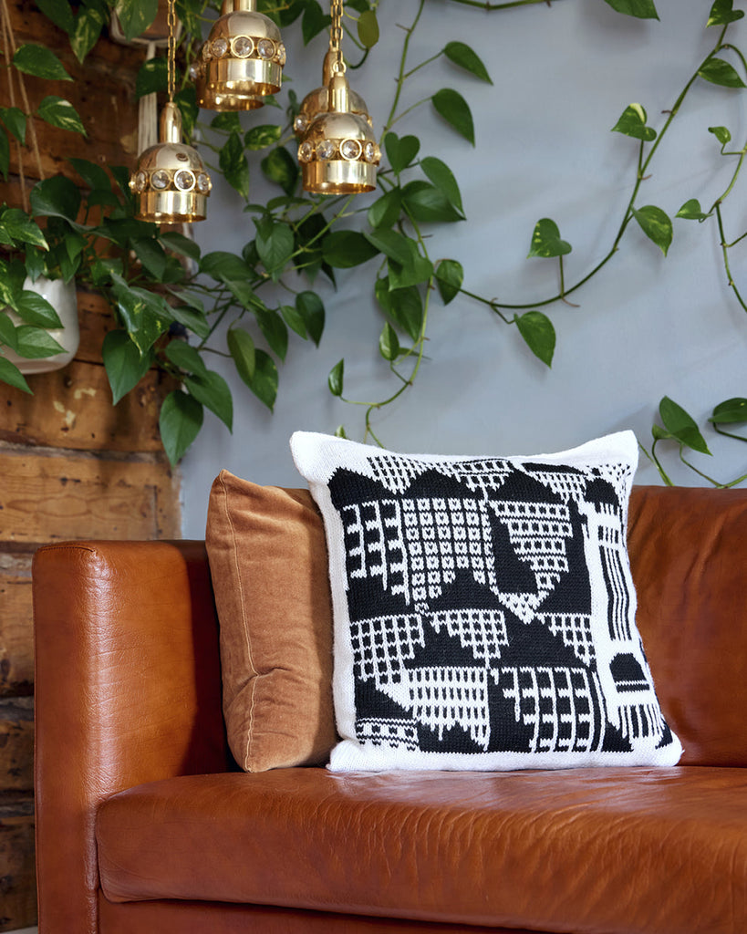 Decorative pillow on a brown couch with greenery and gold pendant lights in the background