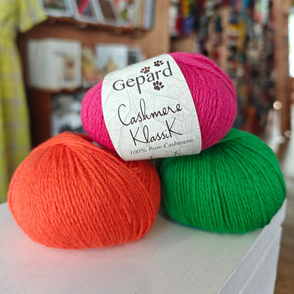 Three skeins of Gepard Cashmere Klassik yarn in orange, pink, and green colors, displayed on a surface.