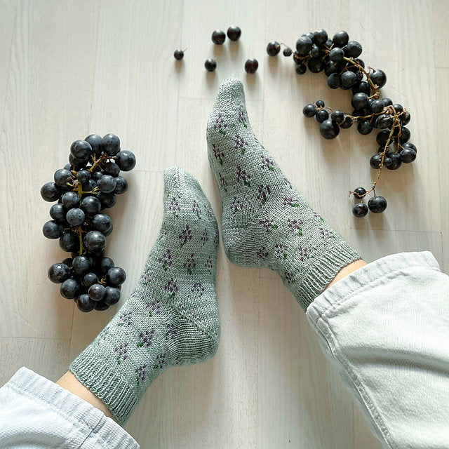 Green floral-patterned socks worn on a light wooden floor with clusters of dark grapes.