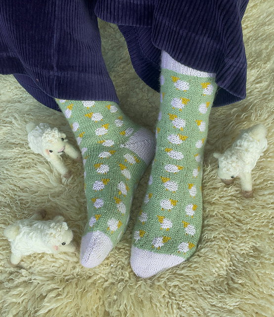 Green socks with white and yellow patterns worn by a person on a fluffy surface with small stuffed animals.