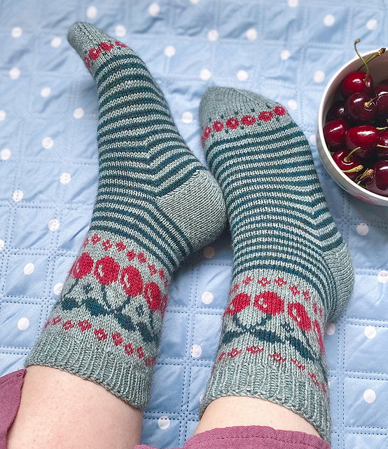 Pair of green and red patterned socks on feet with a bowl of cherries on a polka dot blanket.