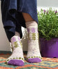 Person wearing white socks with green leaf patterns and purple accents, standing next to a potted plant.