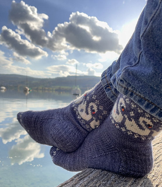 Person wearing patterned socks by a lake with boats and mountains in the background