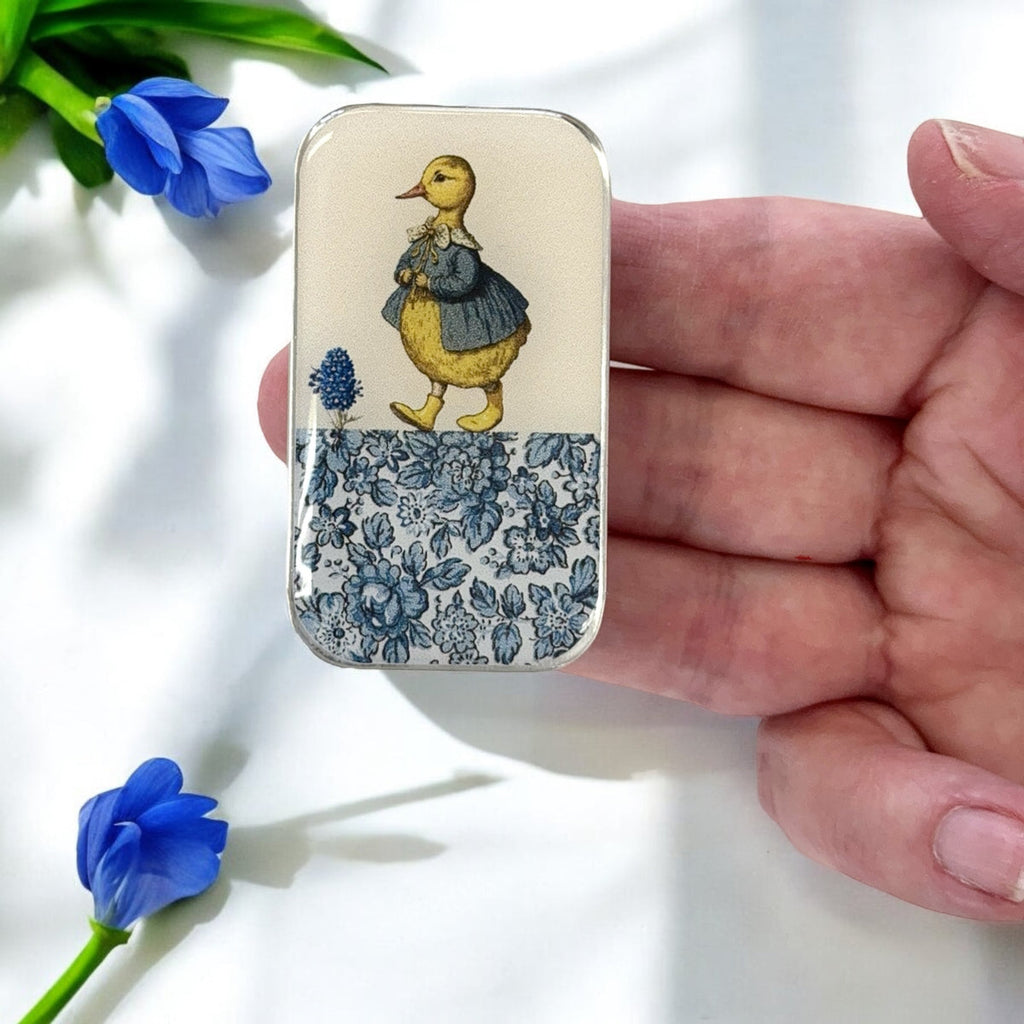 Small tin with a duck illustration held by a hand, with blue flowers in the background