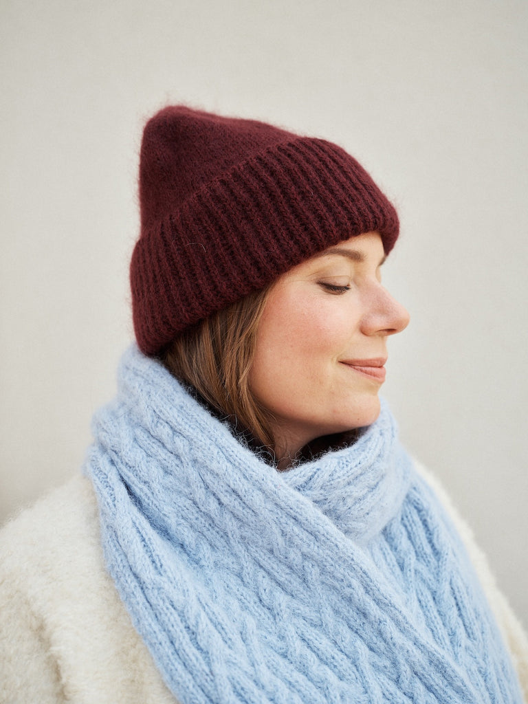Woman wearing a burgundy knit beanie and light blue scarf against a plain background