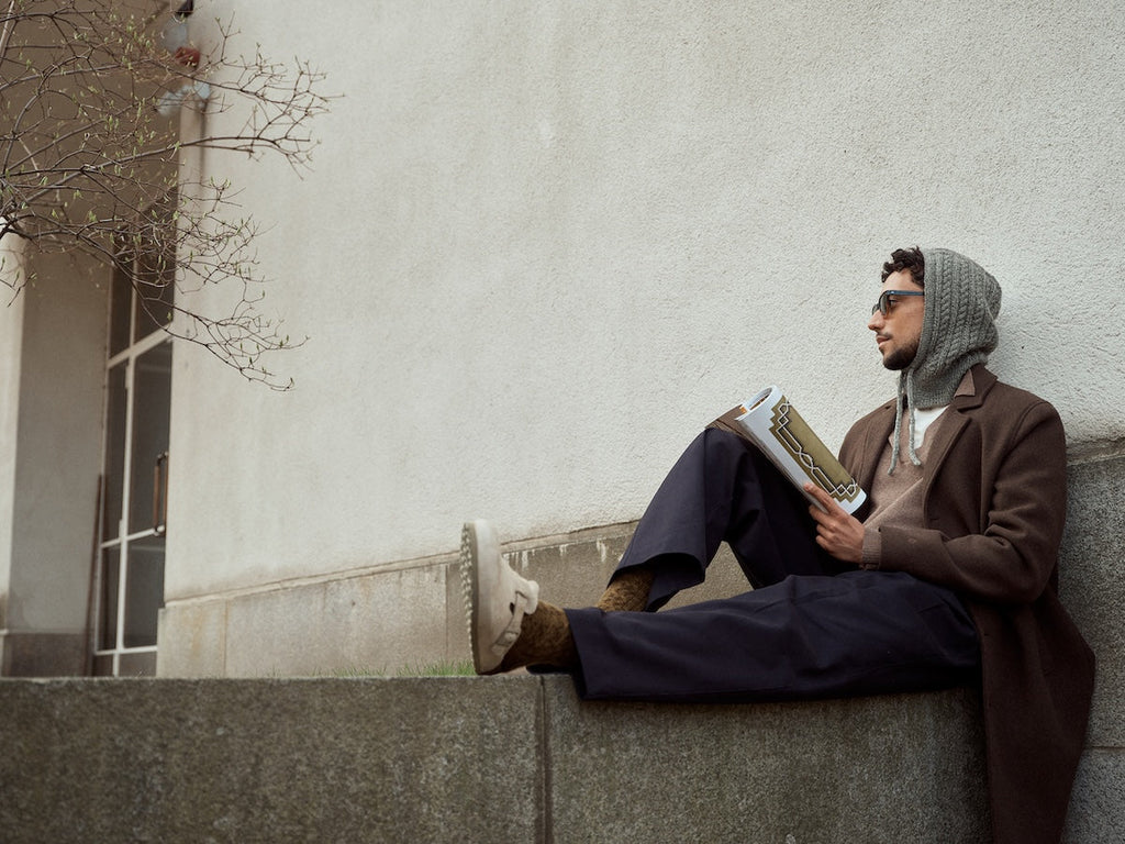 Man sitting on a ledge reading a book against a plain wall.