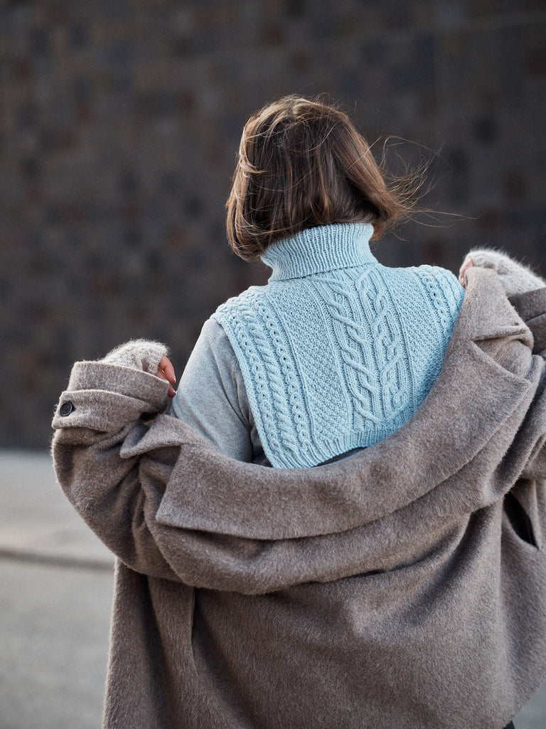 Person wearing a light blue knitted sweater with a brown coat over it, standing against a dark background.