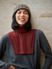 Person wearing a gray sweater with a red knitted turtleneck and a knit beanie against a textured wall.