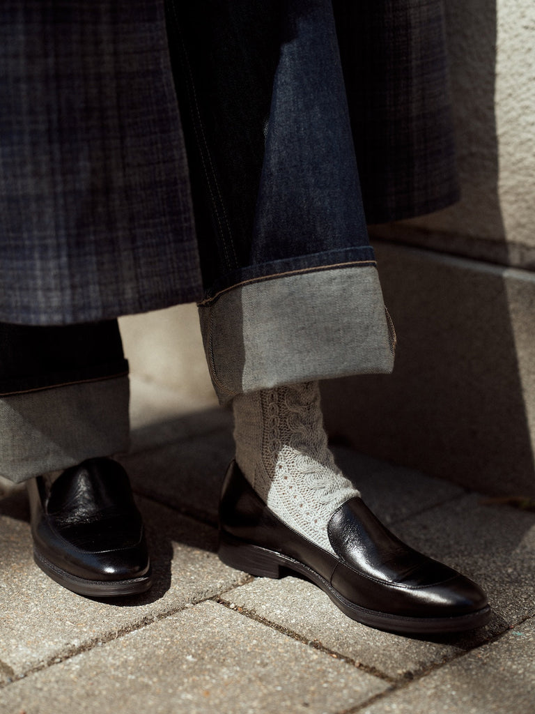 Person wearing black shoes, rolled-up jeans, and a checkered coat on a pavement.