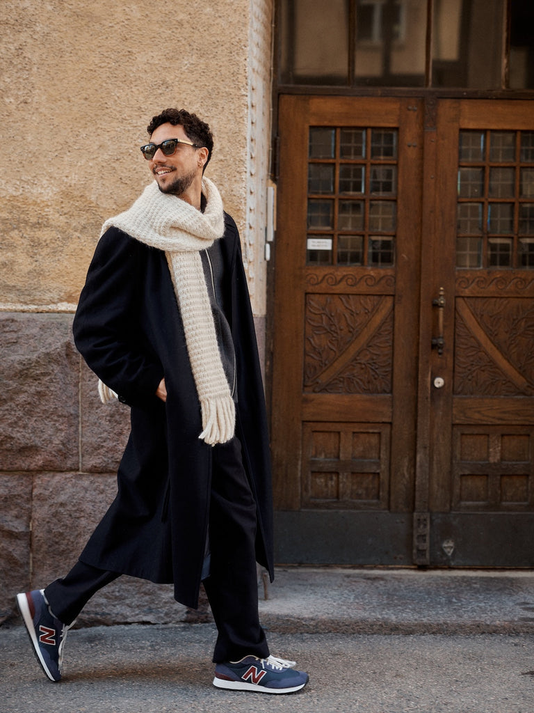 Man wearing a long coat, scarf, and sunglasses walking on a street.