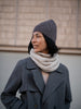 Person wearing a gray knit hat and coat with a beige scarf against a neutral background