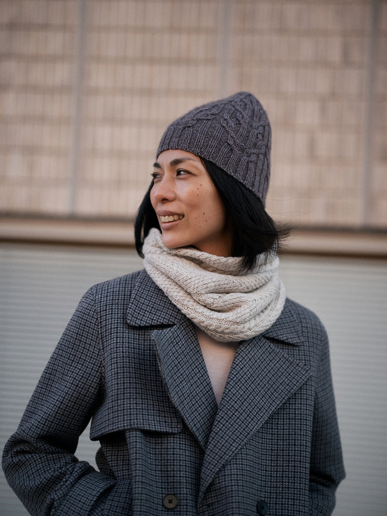 Person wearing a gray knit hat and coat with a beige scarf against a neutral background