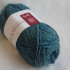 A skein of Jamieson & Smith yarn in a heathered teal blue color, wrapped with a white paper label featuring a red logo block.