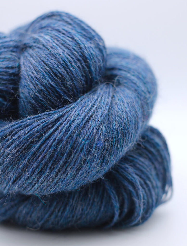 A close-up of a skein of textured yarn in a heathered medium-to-dark blue color, showing the fuzzy, twisted fibers against a clean white background.