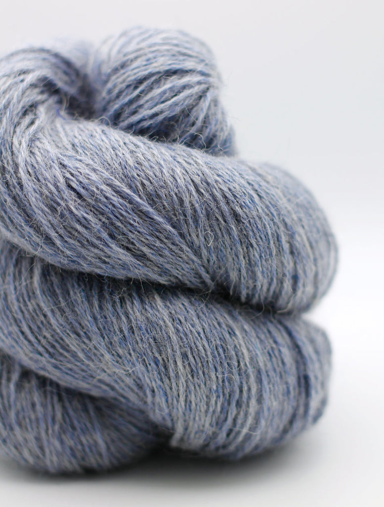 A close-up of a twisted skein of fuzzy yarn in a heathered dusty blue-gray color, set against a clean white background.