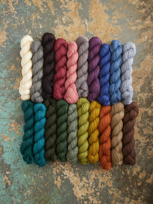 Twisted yarn skeins arranged in two rows on a distressed, textured teal and brown background. The top row includes cream, charcoal, black, dusty rose, taupe, lavender, cobalt, and light blue.