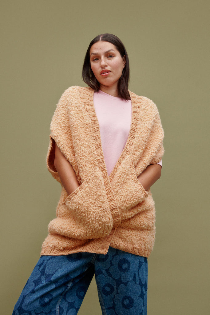 Woman wearing a beige knitted poncho over a pink top and blue patterned pants on a green background