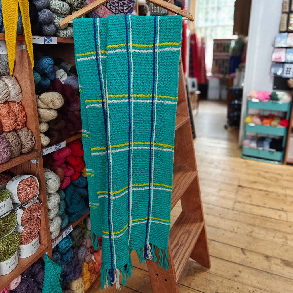 Teal plaid scarf with yellow and white stripes hanging on a wooden rack in a yarn store.