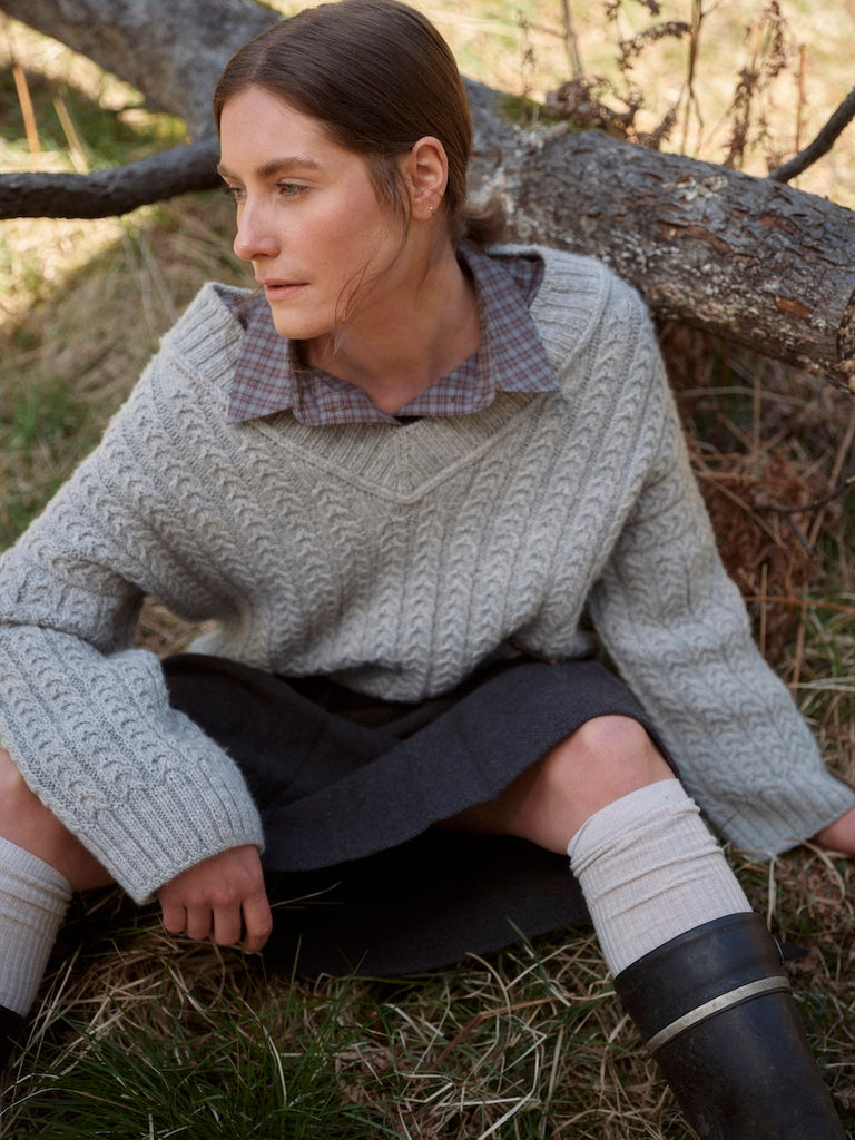 Woman sitting outdoors wearing a cable knit sweater and dark pants.