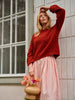 Woman wearing a red knitted sweater and pink skirt holding a woven bag with flowers, standing against a tiled wall.