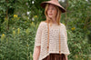 Woman wearing a beige crochet top and wide-brimmed hat in a natural setting with greenery.