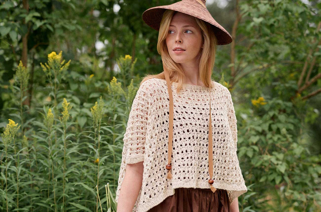 Woman wearing a beige crochet top and wide-brimmed hat in a natural setting with greenery.
