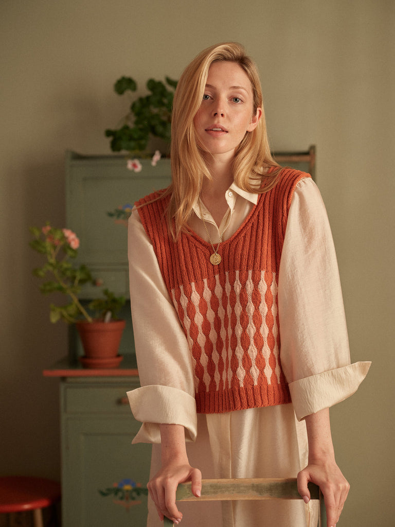 Woman wearing a patterned vest over a white shirt in a room with plants and furniture.