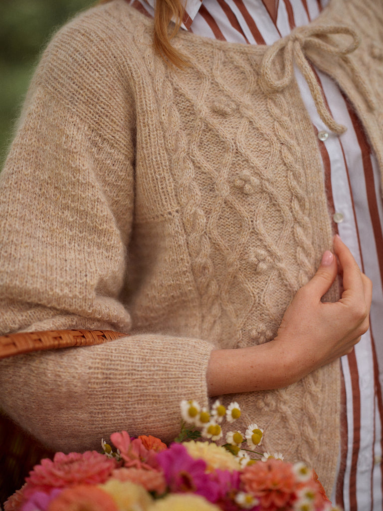 Person wearing a beige cable knit sweater with a blurred background