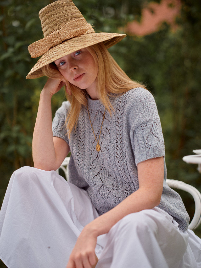 Woman wearing a straw hat and light gray knit top sitting outdoors.