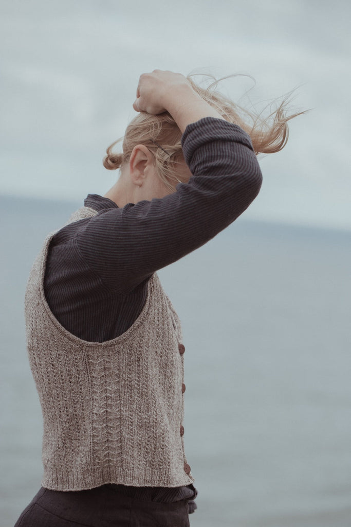 Person wearing a brown sweater and beige vest against a blurred natural background