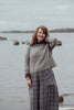 Woman wearing a gray sweater and plaid pants standing by a body of water.