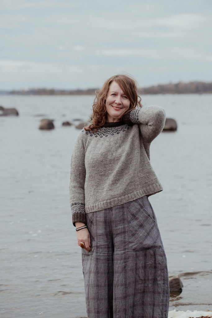Woman wearing a gray sweater and plaid pants standing by a body of water.