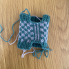 A small handmade knitted pouch or bag in a teal and white fair isle pattern, laid flat on a light wood grain surface.