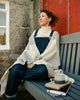 Woman sitting on a bench with a white knit shawl, wearing overalls, in front of a red door.