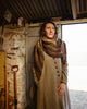 Woman wearing a patterned shawl and long coat standing in a rustic setting.