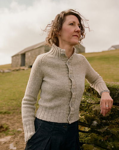 Woman wearing a beige knitted sweater standing outdoors with a stone building and grassy field in the background.