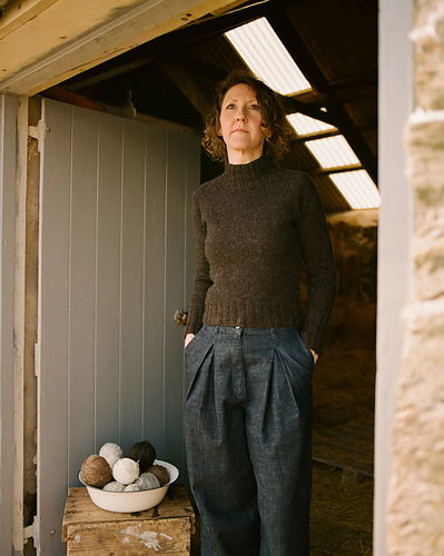 Woman standing in a rustic interior setting with wooden beams and a bowl of yarn.