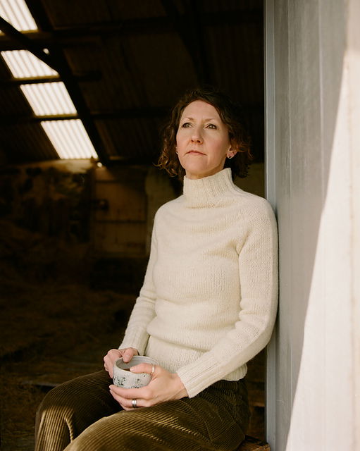 Woman in a white sweater holding a cup, leaning against a wall indoors.