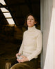 Woman in a white sweater holding a cup, leaning against a wall indoors.