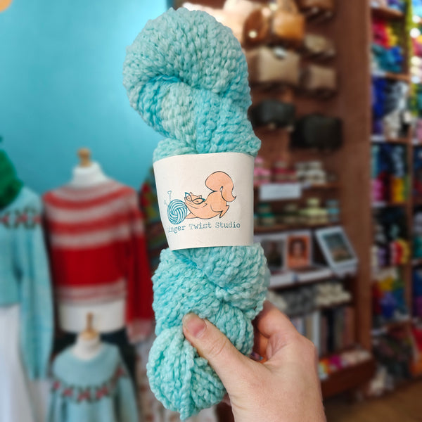 Turquoise yarn skein held in a hand with a blurred store background