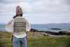 A woman viewed from behind stands on a grassy coastal hill wearing a cream blouse with dramatic puffed sleeves, a multicolor geometric knit vest, and a knit bonnet, looking out over the sea and rocks under a cloudy sky.