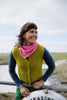 A woman outdoors wearing a bright yellow-green knit vest over a dark blue long-sleeve top and a pink striped knit scarf, leaning on a metal railing with a scenic coastal background.