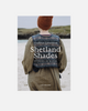 Book cover for 'Shetland Shades: Stories in Wool' by Gudrun Johnston, published by Lane Publishing. The cover features a photograph of a person from behind wearing a brown knit beanie and a patterned blue-and-white wool sweater vest over an olive green dress, standing on a beach.