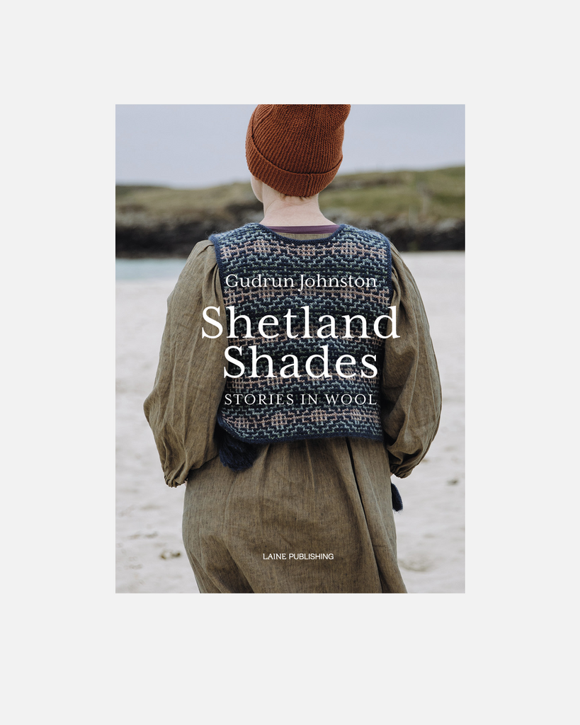 Book cover for 'Shetland Shades: Stories in Wool' by Gudrun Johnston, published by Lane Publishing. The cover features a photograph of a person from behind wearing a brown knit beanie and a patterned blue-and-white wool sweater vest over an olive green dress, standing on a beach.