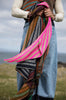 A person outdoors holding a large, vibrant hand-knitted shawl with bright pink, green, brown, and black patterned stripes.