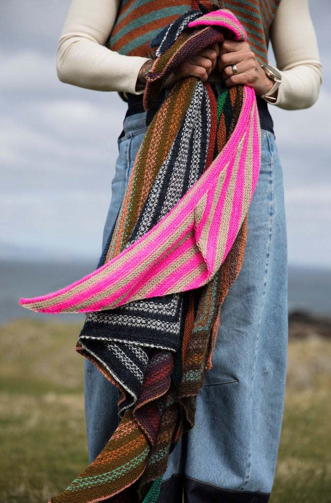 A person outdoors holding a large, vibrant hand-knitted shawl with bright pink, green, brown, and black patterned stripes.