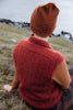 A person sitting outdoors on grassy ground, shown from behind, wearing a rust-orange knit beanie, a matching textured knit sweater vest over a rust-orange long-sleeve top, and plaid pants. The background shows a blurred natural landscape with water and rocks.