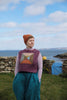 A woman standing outdoors by the sea wearing a textured geometric patterned sweater vest over a light pink long-sleeve top and teal wide-leg trousers, with an orange knit beanie. The background features a stone wall, grass, a blue gate, and a cloudy blue sky.